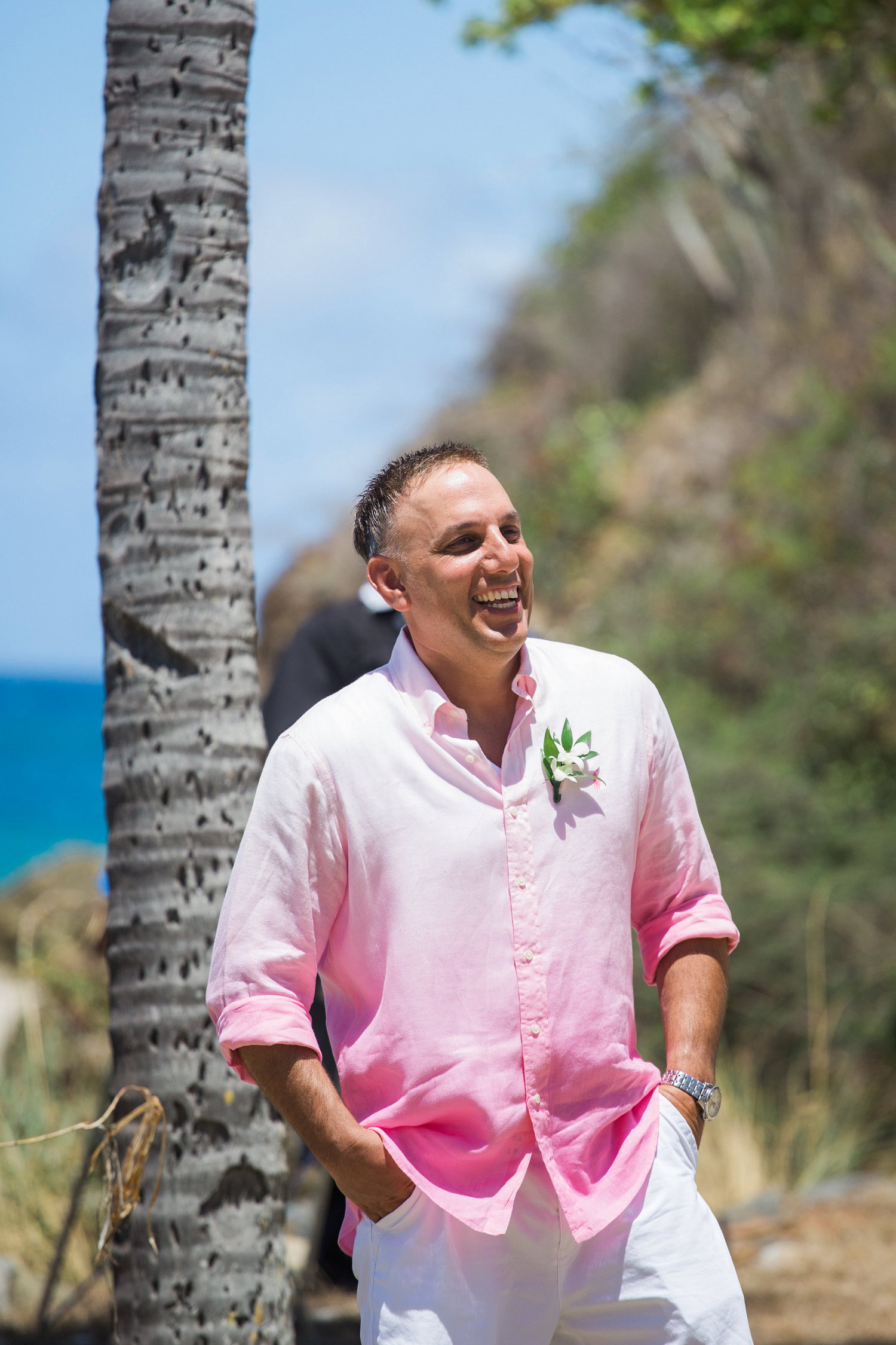 Smiling groom in pink shirt with white orchid boutonniere sees the bride for the first time at vow renewal ceremony.